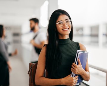 Eine junge Frau mit langen, schwarzen Haaren und Brille hält Bücher in der Hand und lächelt, während sie in einem modernen Lernumfeld steht.
