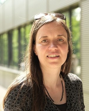 Eine Frau mit langen, braunen Haaren und einer schwarzen Bluse lächelt in die Kamera, während sie draußen steht. Sonnenbrille auf dem Kopf.
