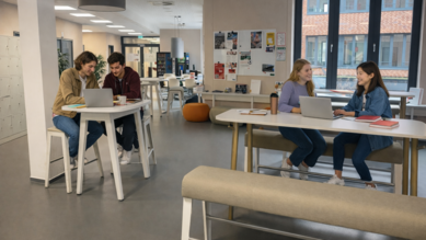 Zwei Gruppen von Studierenden arbeiten an Laptops in einem modernen Lernraum. Tische und Stühle sind hell und einladend gestaltet.