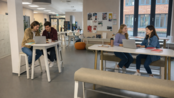 Zwei Gruppen von Studierenden arbeiten an Laptops in einem modernen Lernraum. Tische und Stühle sind hell und einladend gestaltet.