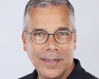 Ein Mann mit kurzen grauen Haaren und Brille lächelt in die Kamera. Er trägt ein schwarzes Polohemd und hat einen neutralen Gesichtsausdruck. Der Hintergrund ist hell und einfarbig.