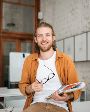 Ein junger Mann mit langen Haaren sitzt an einem Tisch in einem modernen Raum. Er hält eine Mappe mit Dokumenten und eine Brille in der Hand. Im Hintergrund sind einige Bilder an der Wand zu sehen.