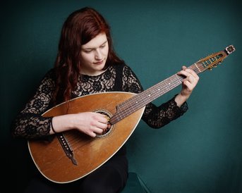 Eine Frau mit langen, roten Haaren spielt eine Mandoline, während sie auf einem grünen Hintergrund sitzt. Sie trägt ein schwarzes, spitzenbesetztes Oberteil.