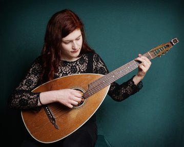 Eine Frau mit langen, roten Haaren spielt eine Mandoline, während sie auf einem grünen Hintergrund sitzt. Sie trägt ein schwarzes, spitzenbesetztes Oberteil.