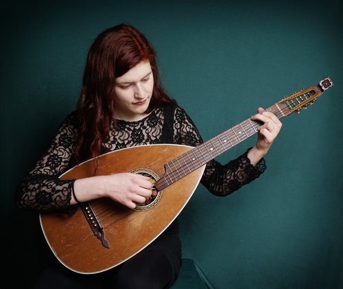 Eine Frau mit langen, roten Haaren spielt eine Mandoline, während sie auf einem grünen Hintergrund sitzt. Sie trägt ein schwarzes, spitzenbesetztes Oberteil.
