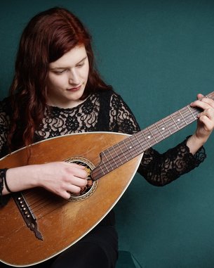 Eine Frau mit langen, roten Haaren spielt eine Mandoline, während sie auf einem grünen Hintergrund sitzt. Sie trägt ein schwarzes, spitzenbesetztes Oberteil.