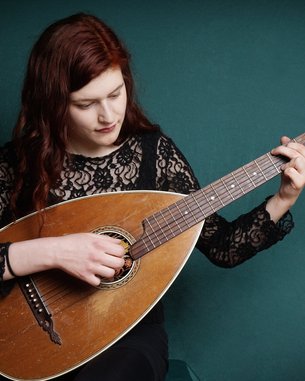 A woman with long, red hair is playing a mandolin while sitting against a green background. She is wearing a black top with lace detailing.