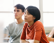 Three students are sitting in a classroom, listening attentively. A woman with short black hair is wearing an orange top, while two men next to her are dressed in neutral colors.