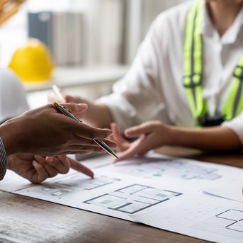 Two people are discussing construction plans at a table while holding pens and blueprints. Safety helmets are visible.