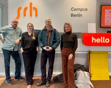 Four people are standing at the reception of the SRH Campus Berlin, with a 'hello' sign in the background.