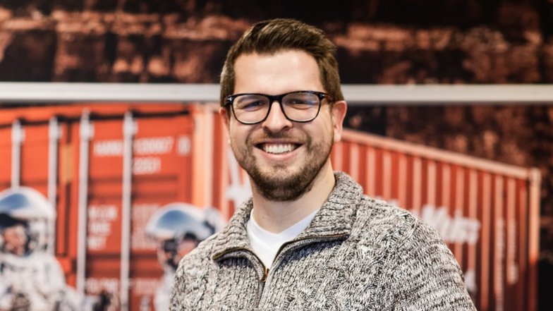 Ein Mann mit Brille und Bart lächelt in einem grauen Pullover vor einem Hintergrund mit Containern und Raumfahrern.
