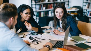 Three students are working at a table, surrounded by notes and laptops, as they discuss a project together.