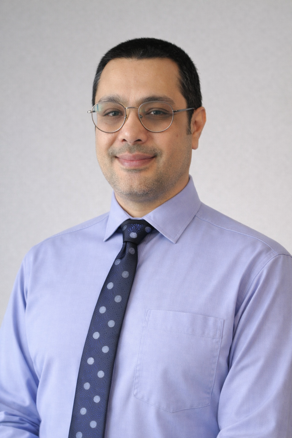 A man with glasses is wearing a light blue shirt and a dark blue polka-dotted tie. He is smiling slightly against a neutral background.