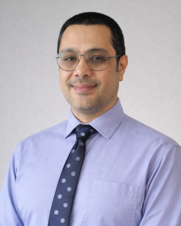 A man with glasses is wearing a light blue shirt and a dark blue polka-dotted tie. He is smiling slightly against a neutral background.
