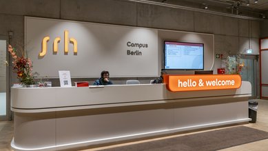 Empfangstresen des SRH Campus Berlin mit einer Mitarbeiterin, einem Bildschirm und der Begrüßung "hello & welcome". Blumenarrangements sind dekorativ platziert.