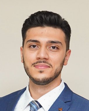 Ein junger Mann mit dunklem Haar trägt einen blauen Anzug und ein weißes Hemd mit Punkten. Er blickt direkt in die Kamera.