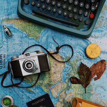 An old camera, a United States passport, a yellow folder, and various items are laid out on a world map. Some sheets of paper and a compass needle are also visible, hinting at the theme of travel.