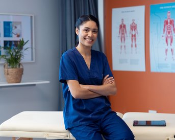 A young woman in blue work attire is sitting on a treatment table and smiling. In the background, anatomical diagrams and a plant are visible.