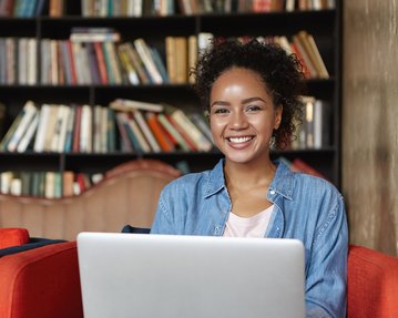 Eine lächelnde Frau mit lockigem Haar sitzt auf einem roten Sofa und arbeitet an einem Laptop. Im Hintergrund sind Bücherregale mit einer Vielzahl von Büchern sichtbar. Die Atmosphäre wirkt gemütlich und einladend.