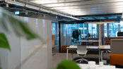 A modern office with bright walls, tables, and chairs. In the background, large windows and plants are visible. The space feels open and inviting.