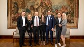 Eine Gruppe von sieben Personen steht lächelnd vor einem Wandteppich, gekleidet in formelle Kleidung, in einem eleganten Raum.