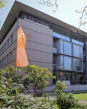 Das Gebäude mit großen Fenstern und einem modernen Design trägt das Logo "sitch" an der Fassade. Vor dem Gebäude sind grüne Pflanzen und ein orangefarbener Fahne sichtbar.