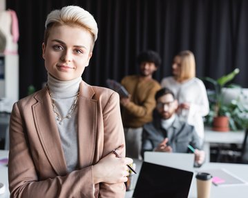 A young woman with blonde hair and a blazer stands in the foreground, while three people in the background are working at a table.