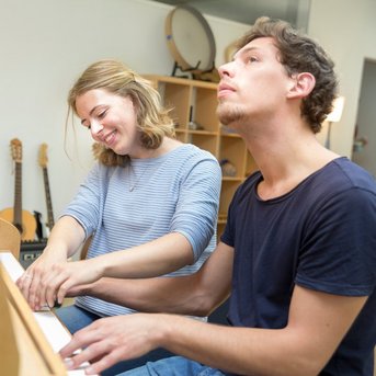 Zwei Personen sitzen an einem Klavier, während sie zusammen spielen. Die Frau lächelt, während der Mann konzentriert schaut. Im Hintergrund sind Musikinstrumente sichtbar.