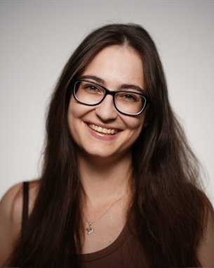 Eine lächelnde Frau mit langen, dunklen Haaren und Brille, die in die Kamera schaut. Sie trägt ein einfaches, ärmelloses Oberteil.