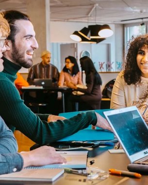 A group of five people is sitting at a table, engaging in lively discussion and smiling. On the table are notebooks, a laptop, and various writing utensils. In the background, more people can be seen.