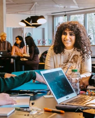 Eine Gruppe von fünf Personen sitzt an einem Tisch, diskutiert lebhaft und lächelt. Auf dem Tisch liegen Notizbücher, ein Laptop und verschiedene Schreibutensilien. Im Hintergrund sind weitere Personen zu sehen.