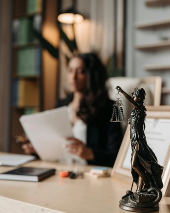 Eine Frau sitzt an einem Schreibtisch, hält Dokumente in der Hand. Im Vordergrund steht eine Statue der Justitia mit Waage. Der Hintergrund zeigt eine moderne Büroumgebung mit Regalen und Pflanzen.