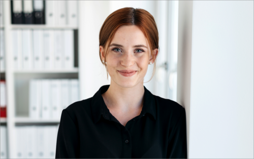 Eine junge Frau mit roten Haaren lächelt freundlich in einem modernen Büro, umgeben von Aktenordnern. Sie trägt ein schwarzes Hemd und steht an einer Wand.