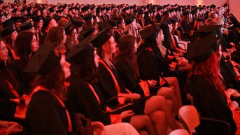 A large group of graduates in academic gowns and caps is seated in a festive room during a graduation ceremony.