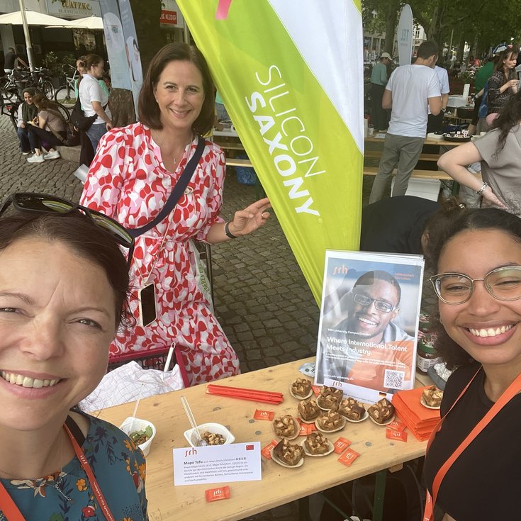 Three women are smiling as they pose in front of a food stall with a sign. In the background, more people and stalls can be seen.