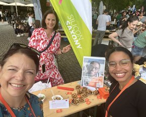 Three women are smiling as they pose in front of a food stall with a sign. In the background, more people and stalls can be seen.