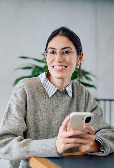 A young woman with glasses sits at a table, holding a smartphone in her hand, smiling and dressed in a cozy sweater.