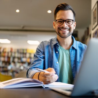 A smiling man sits at a table in a café, working on a laptop and holding a pen in his hand. In the background, bookshelves are visible.