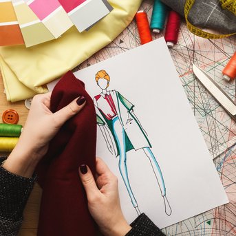 Hands are holding a fabric, and a fashion sketch lies on a table surrounded by sewing supplies and color swatches.