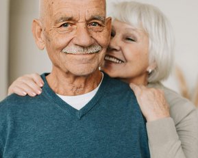 An older man in a gray sweater smiles at the camera while an older woman with gray hair gently wraps her arms around his shoulders from behind. Both appear relaxed and friendly.