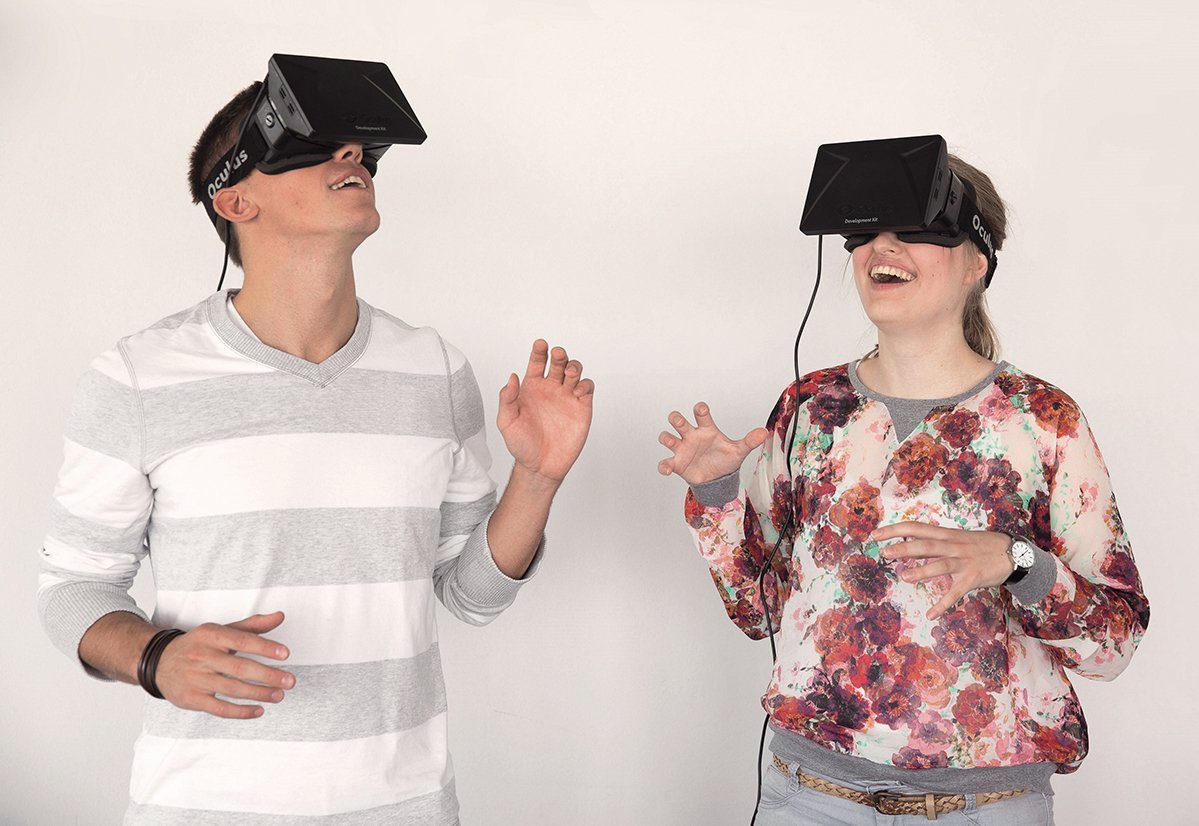 Two people are wearing virtual reality headsets and interacting with each other. The man is wearing a striped top, while the woman has on a floral shirt. Both seem to be immersed in a VR experience.