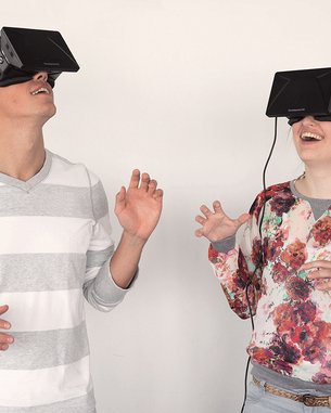 Zwei Personen tragen Virtual-Reality-Headsets und interagieren miteinander. Der Mann trägt ein gestreiftes Oberteil, während die Frau ein blumiges Shirt hat. Beide scheinen in ein VR-Erlebnis vertieft zu sein.