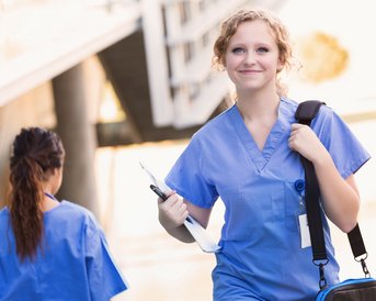 Eine lächelnde Krankenschwester in blauer Arbeitskleidung trägt eine Tasche und ein Clipboard, während sie auf einem Weg geht. Eine weitere Person in ähnlicher Kleidung ist im Hintergrund zu sehen.