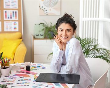 A smiling woman sits at a desk with a graphic tablet, surrounded by color samples and a computer.