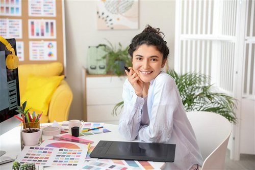 A smiling woman sits at a desk with a graphic tablet, surrounded by color samples and a computer.