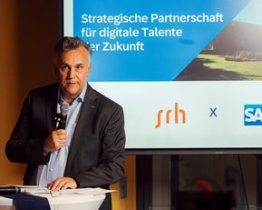 A man stands at a table holding a microphone, while in the background there is a presentation about the strategic partnership between SRH and SAP.