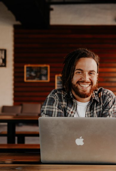A smiling man with long, curly hair is sitting at a table in a café, working on a laptop. Next to him is a glass with a drink.
