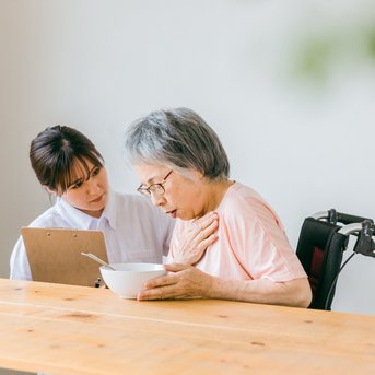 A caregiver is assisting an elderly woman who is holding a bowl of food while sitting at a table.