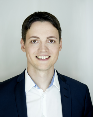 Ein junger Mann mit kurzem, dunklem Haar trägt einen Anzug und lächelt vor einem neutralen Hintergrund.