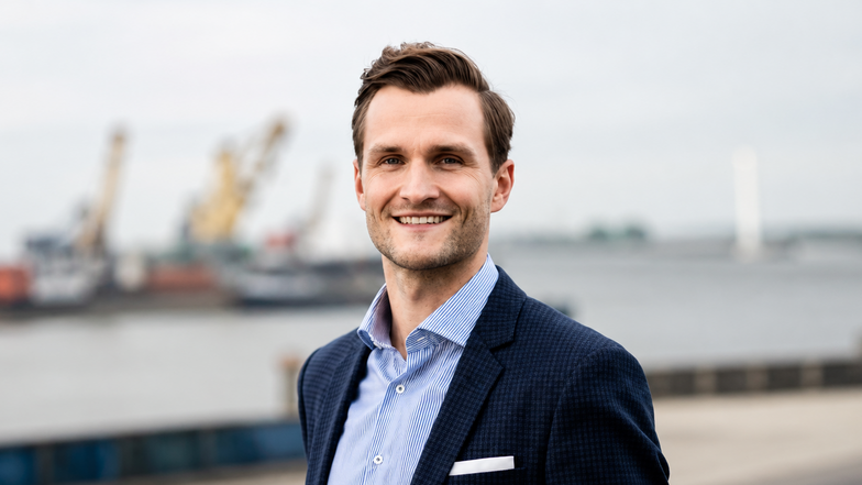 Mann in Anzug steht am Hafen, lächelt in die Kamera, mit Schiffen und Kränen im Hintergrund.
